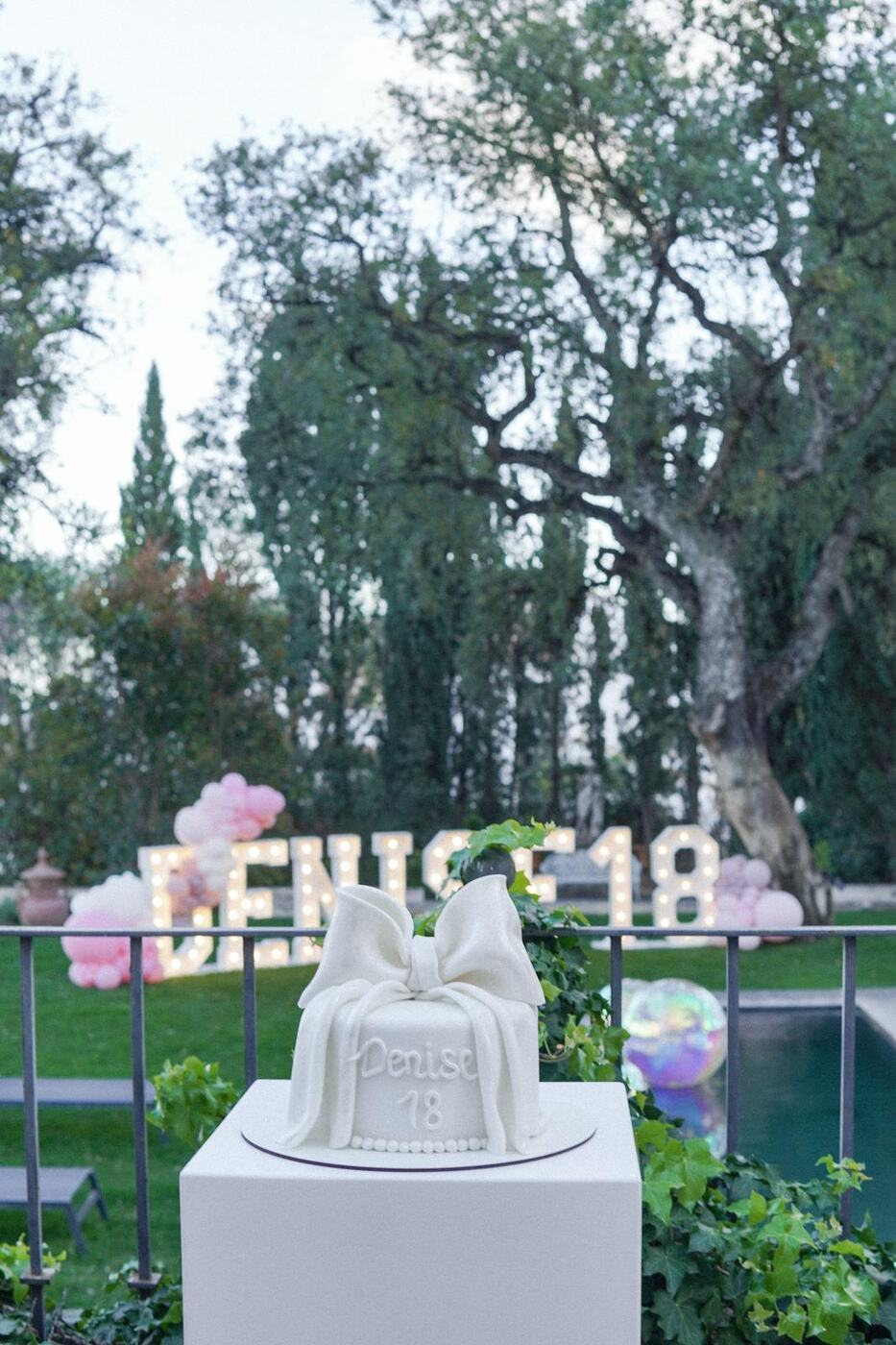 White bow birthday cake in front of light-up letters at dusk, 18th birthday Marbella
